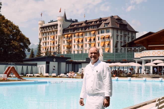 Älterer MAnn in Kochbluse steht vor einem Pool, ein Luxushotel im Hintergrund