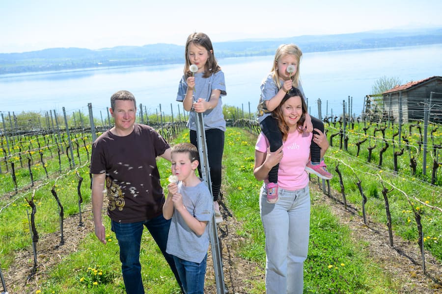 Constantine, 11 avril 2025. Julien et Célia Cressier et leurs trois enfants. En arrière-plan, le lac de Morat. Reportage au Domaine des Marnes. Célia Cressier (t-shirt rose) a repris avec son mari Julien le domaine développé par son père Pierre Gentizon. Le Domaine des Marnes est situé dans l'appellation Vully, localisée entre les lacs de Neuchâtel et de Morat. ©François Wavre | Lundi13