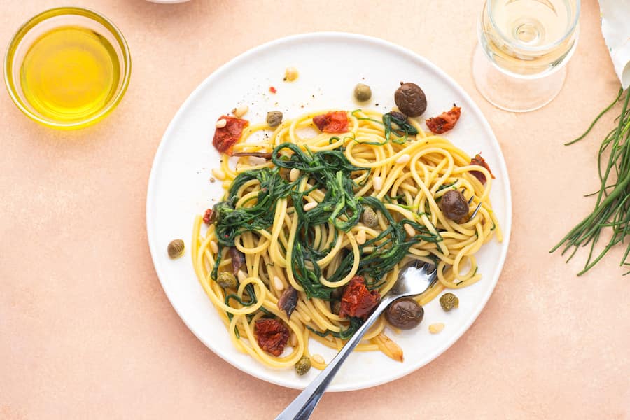 Spaghetti with Italian Agretti, Barba di frate or Saltwort or Salsola Soda, olives, anchovy, tomatoes, capers,  pine nuts and olive oil, spring Italian recipes. Pink soft spring background