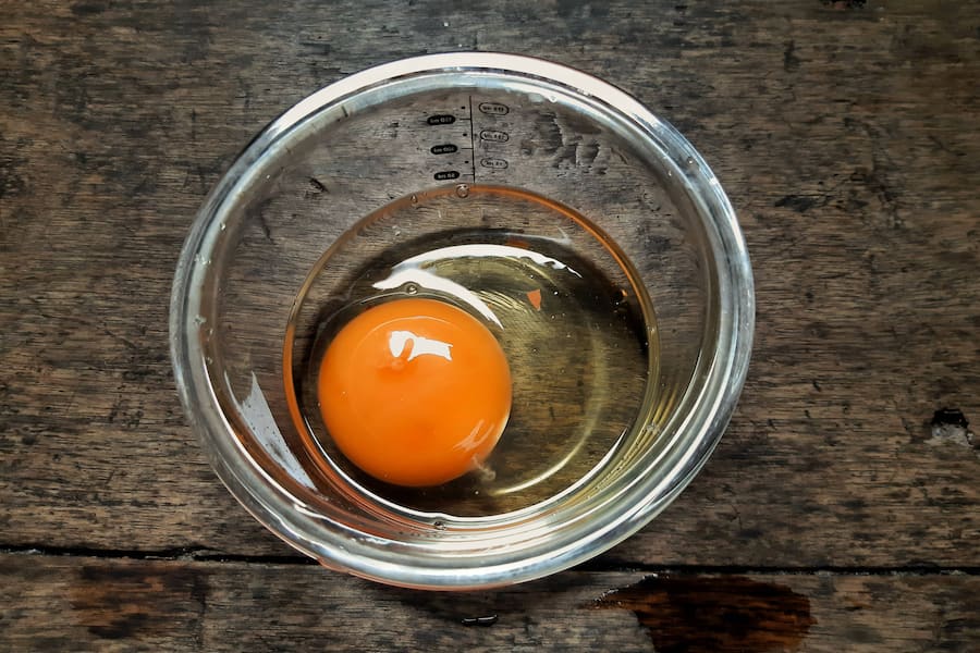 A raw egg with a bright orange yolk sits in a transparent glass bowl on a rustic wooden surface. The reflection on the glass adds depth, highlighting the texture of the wood and the eggâs natural form